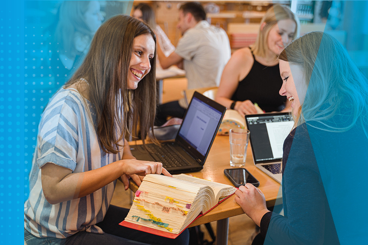 Drei junge Frauen sitzen gemeinsam an einem Arbeitstisch und lernen. Auf dem Tisch befinden sich Laptops, Mobiltelefone und ein aufgeschlagenes Buch.