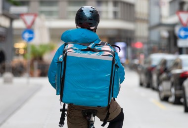 Die Person trägt einen dunklen Helm, sowie eine Jacke und einen Lieferdienst-Rucksack im gleichen hellblauen Farbton. 