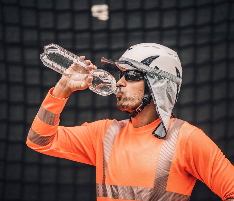 Ein Arbeitnehmer trägt Kleidung zum Schutz gegen Hitze und trinkt Wasser aus einer Flasche.