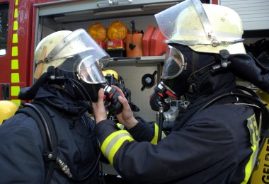 Zwei Feuerwehrleute in vollständiger Schutzausrüstung und Atemschutzmasken stehen vor einem Einsatzfahrzeug. Eine Person hilft der anderen beim Anlegen oder Überprüfen der Atemmaske. Zwei Feuerwehrleute in vollständiger Schutzausrüstung und Atemschutzmasken stehen vor einem Einsatzfahrzeug. Eine Person hilft der anderen beim Anlegen oder Überprüfen der Atemmaske.
