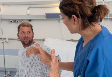 Ein Mann in Krankenhauskleidung sitzt aufrecht im Bett und schaut eine Pflegekraft an, die ihm gegenübersitzt und mit erhobenen Händen eine abwehrende Geste macht. Die Situation wirkt angespannt. Ein Mann in Krankenhauskleidung sitzt aufrecht im Bett und schaut eine Pflegekraft an, die ihm gegenübersitzt und mit erhobenen Händen eine abwehrende Geste macht. Die Situation wirkt angespannt.
