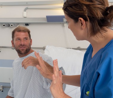 Ein Mann in Krankenhauskleidung sitzt aufrecht im Bett und schaut eine Pflegekraft an, die ihm gegenübersitzt und mit erhobenen Händen eine abwehrende Geste macht. Die Situation wirkt angespannt. Ein Mann in Krankenhauskleidung sitzt aufrecht im Bett und schaut eine Pflegekraft an, die ihm gegenübersitzt und mit erhobenen Händen eine abwehrende Geste macht. Die Situation wirkt angespannt.