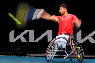 Tennisspieler im Rollstuhl von vorne. Schlägt nach dem Ball, der ausholende Arm ist durch die Bewegung verwachen, der getroffene Ball auch.